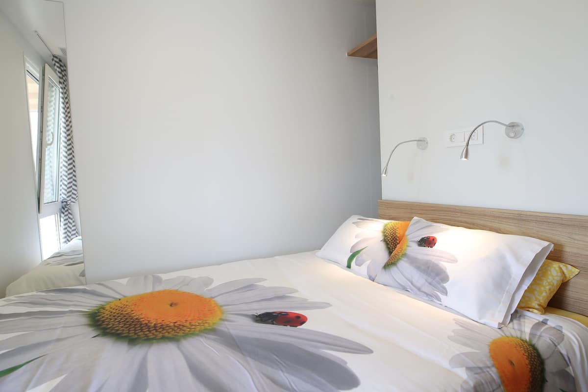 A bedroom with a bed featuring a floral bedspread, a wooden headboard, and a wall-mounted light fixture.