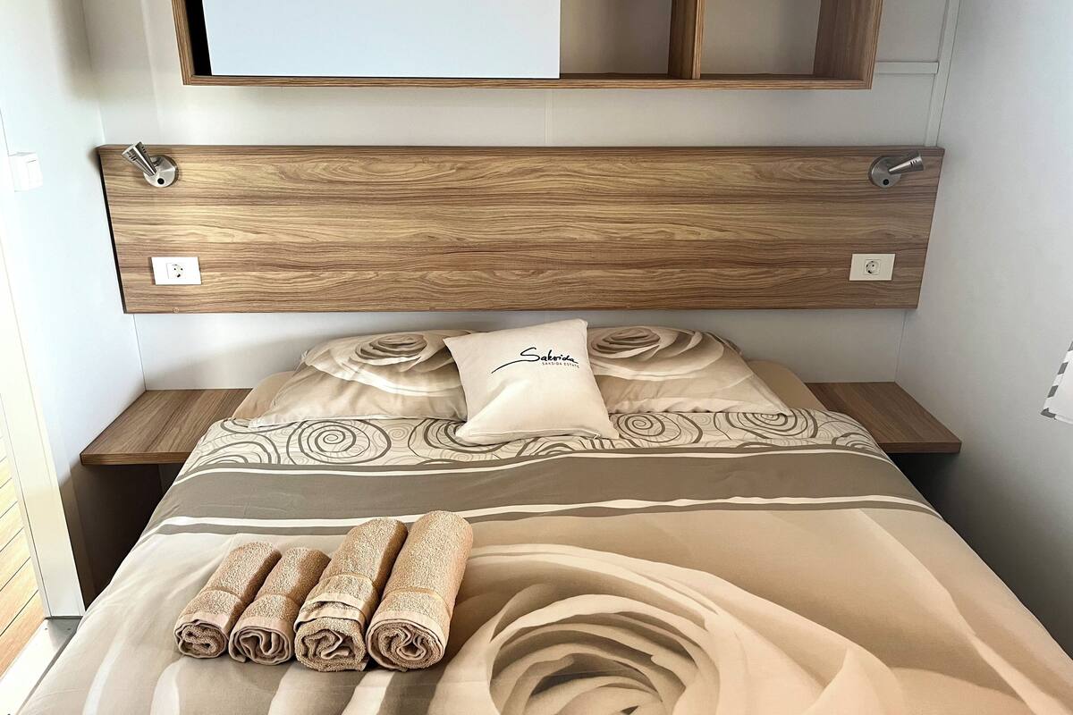 A neatly made bed with a patterned bedspread, a pillow with the text "Sakaria," and rolled towels on the bed.
