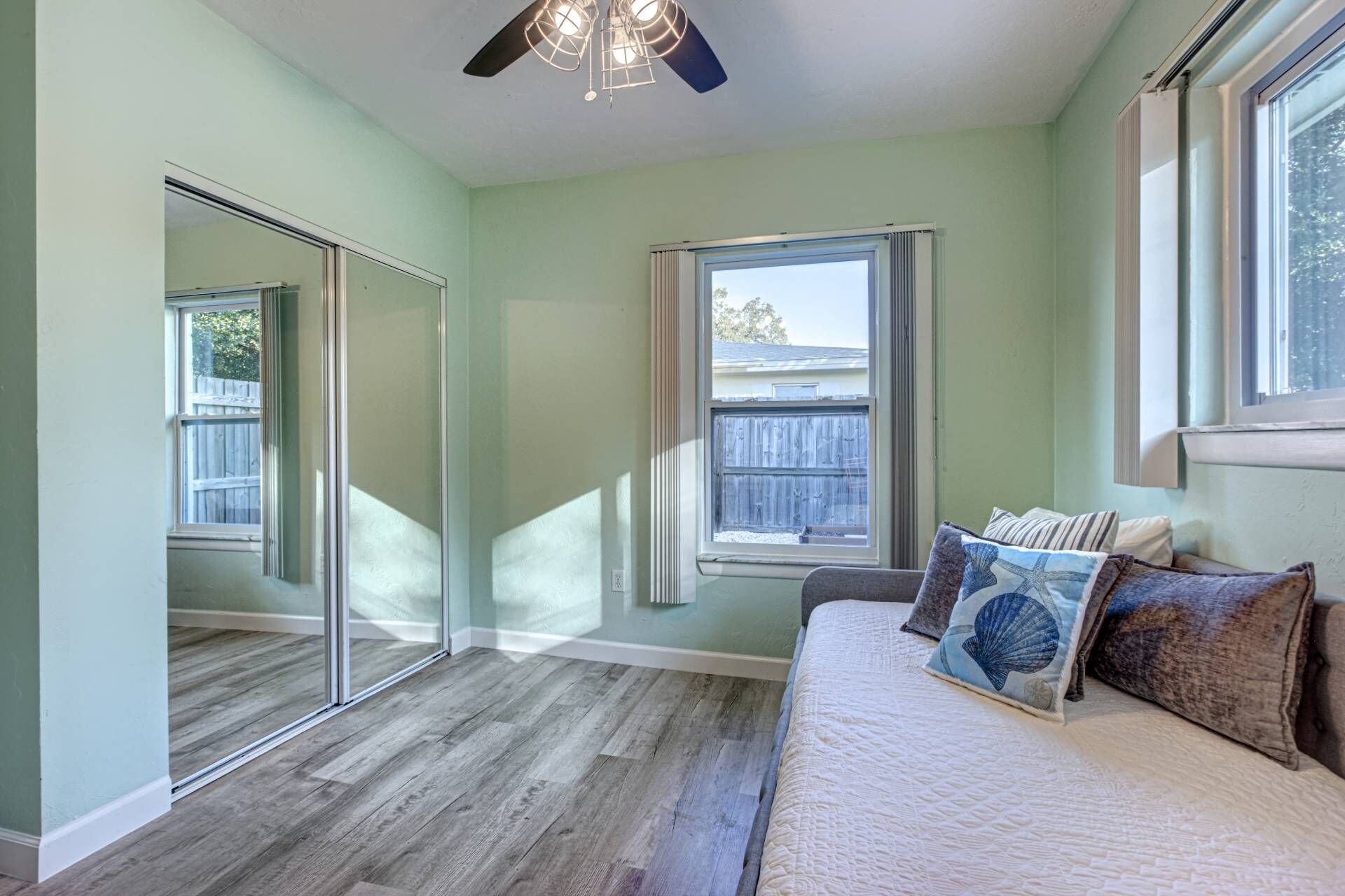 A bedroom with a large mirror, a bed with multiple pillows, a ceiling fan, and a window with a view of a fence.