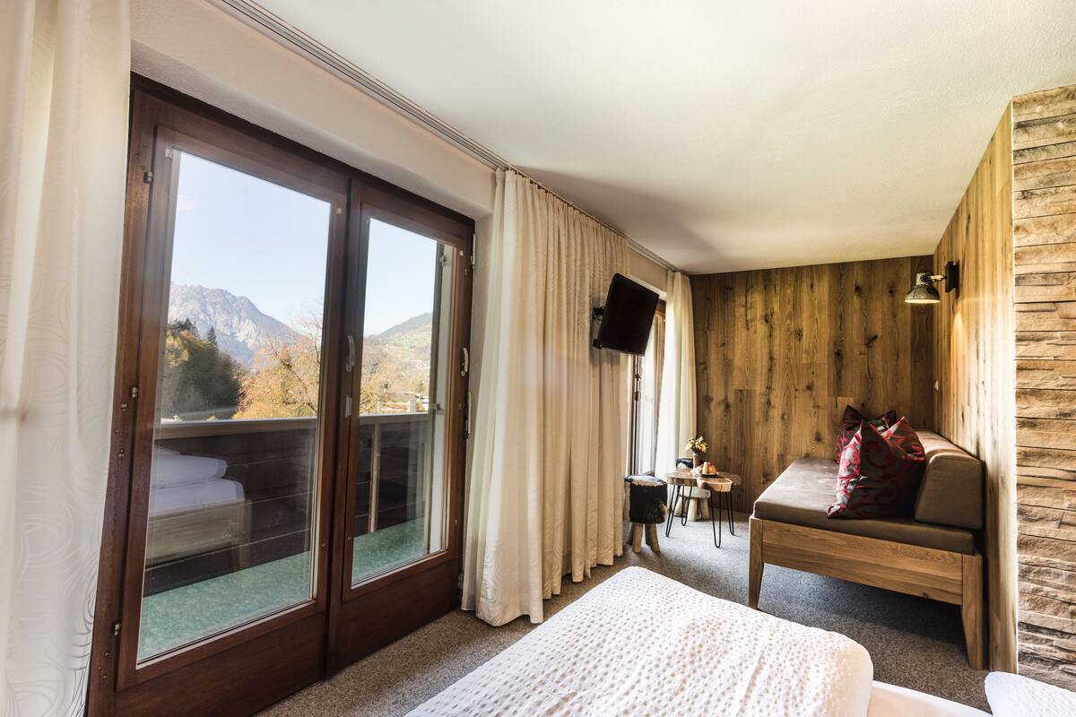A hotel room with a large sliding glass door leading to a balcony, a bed, a wooden bench with cushions, a small table with a chair, and a mounted TV.