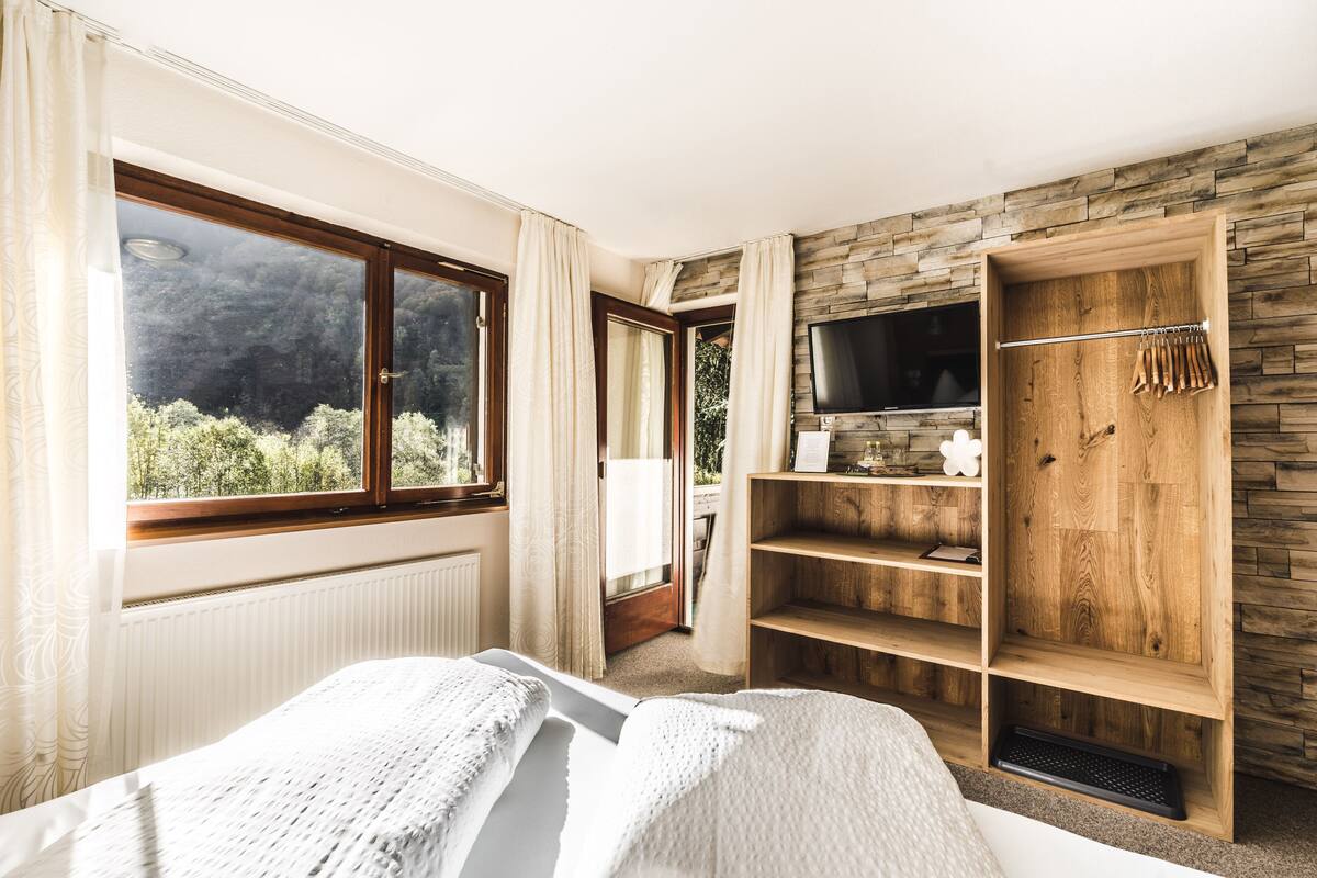 A bedroom with a large window, a bed, a wooden wardrobe with a TV, and a view of greenery outside.