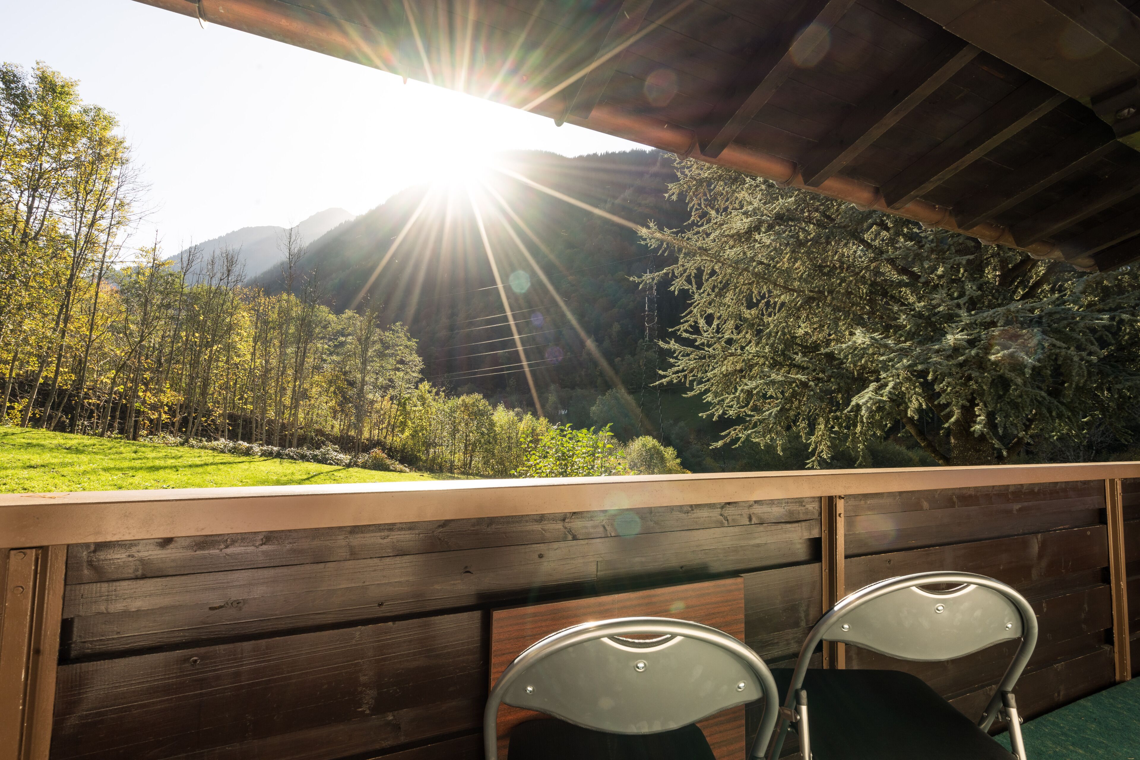 A wooden deck with two metal chairs overlooking a green landscape with trees and mountains.