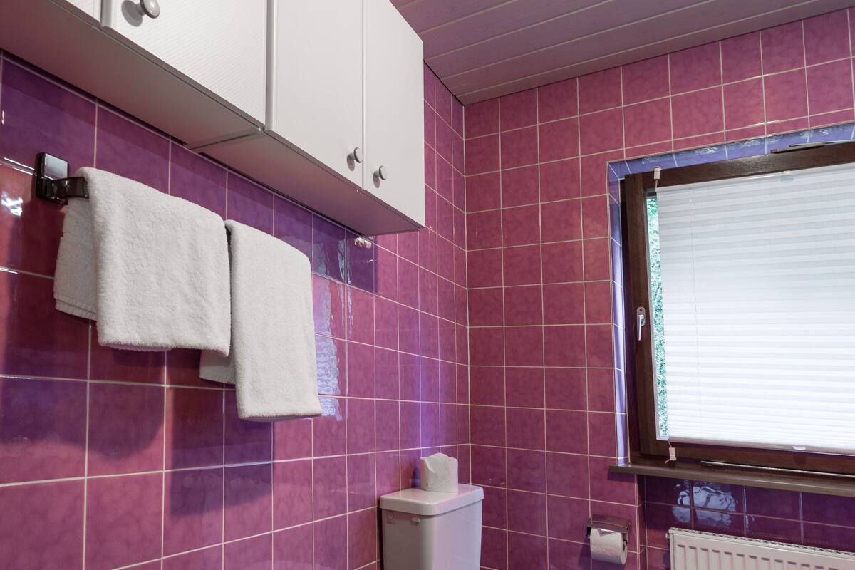 A bathroom with pink tiled walls, a white toilet, a window with blinds, and a black and white patterned floor.