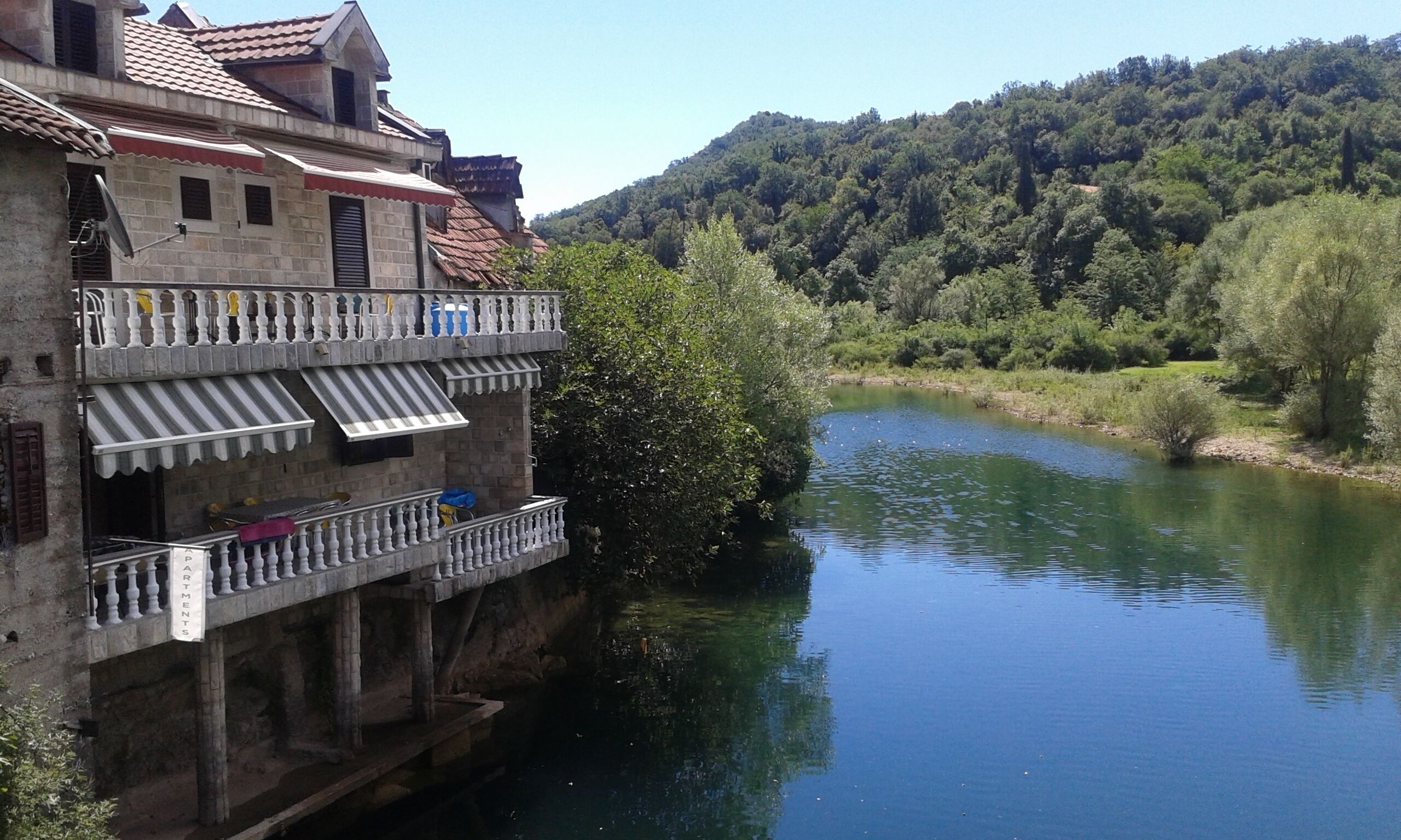 A building with a balcony overlooking a river, surrounded by greenery and hills.