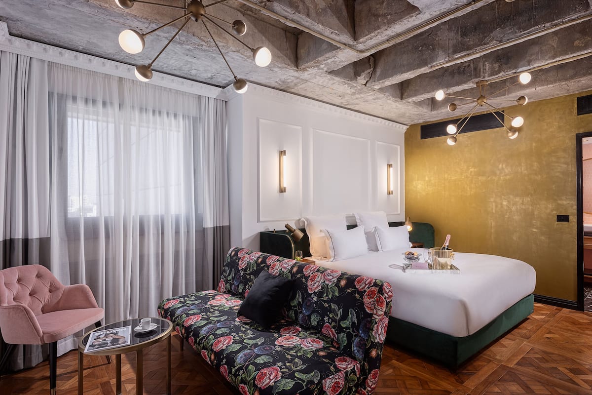 A hotel room with a large bed, a floral sofa, a small table with a cup, and a large window with sheer curtains.