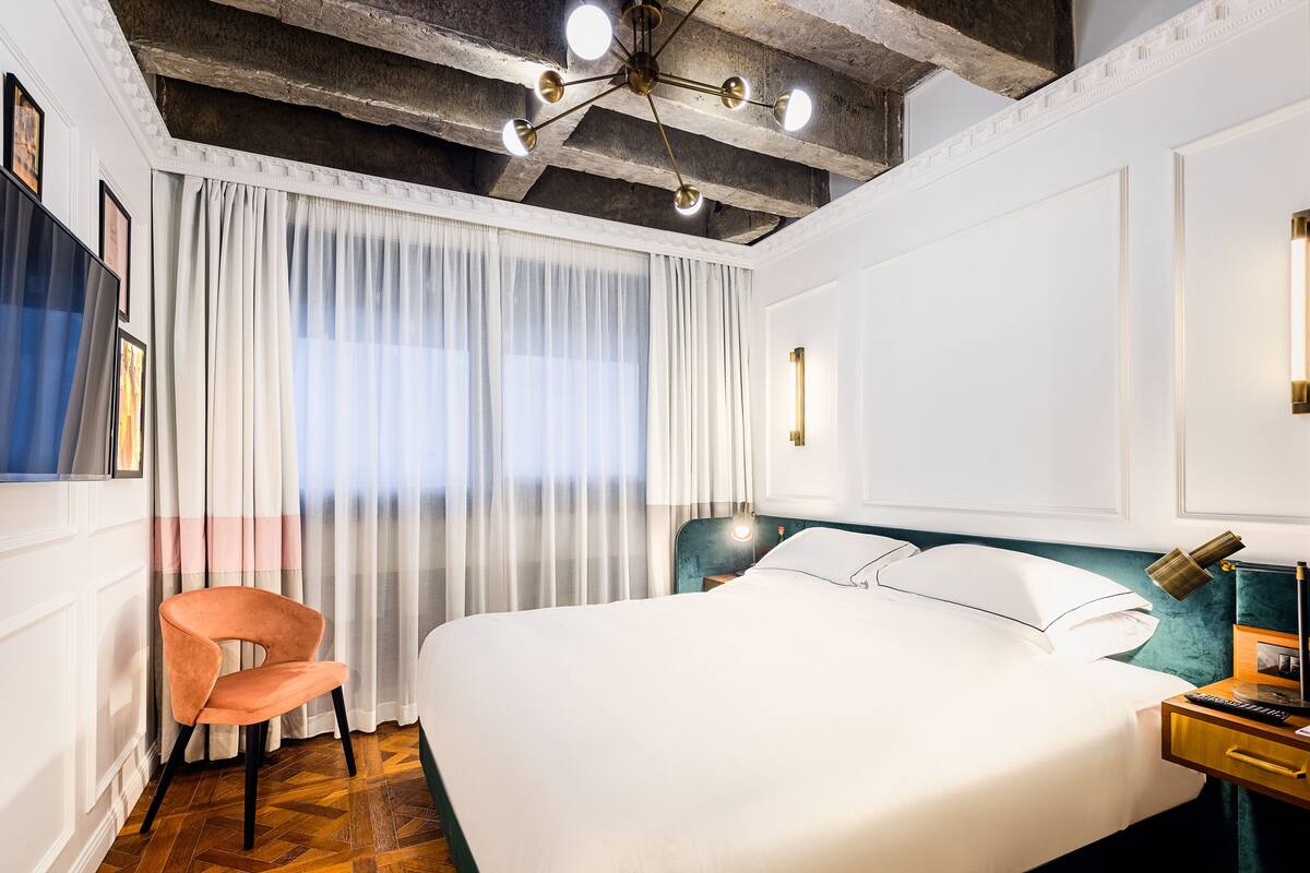 A hotel room with a large bed, a chair, a desk with a lamp, and a ceiling with exposed beams and lights.