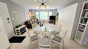 A modern living room with a glass dining table, white chairs, a brown sofa, and a TV. - Thien May Guest House. Up to 6 guests (Surrey)