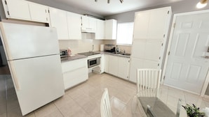 A modern kitchen with white cabinets, a stainless steel refrigerator, a microwave, and a sink. There is a dining area with a glass table and chairs. - Thien May Guest House. Up to 6 guests (Surrey)