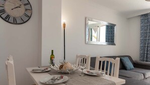 Un coin repas avec une table blanche dressée pour deux personnes, une horloge au mur et un canapé à l’arrière-plan.