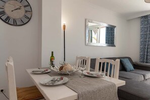 A dining area with a white table set for two, a clock on the wall, and a sofa in the background. - Parque Santiago II 222 - Two Bed (Playa de la Américas)