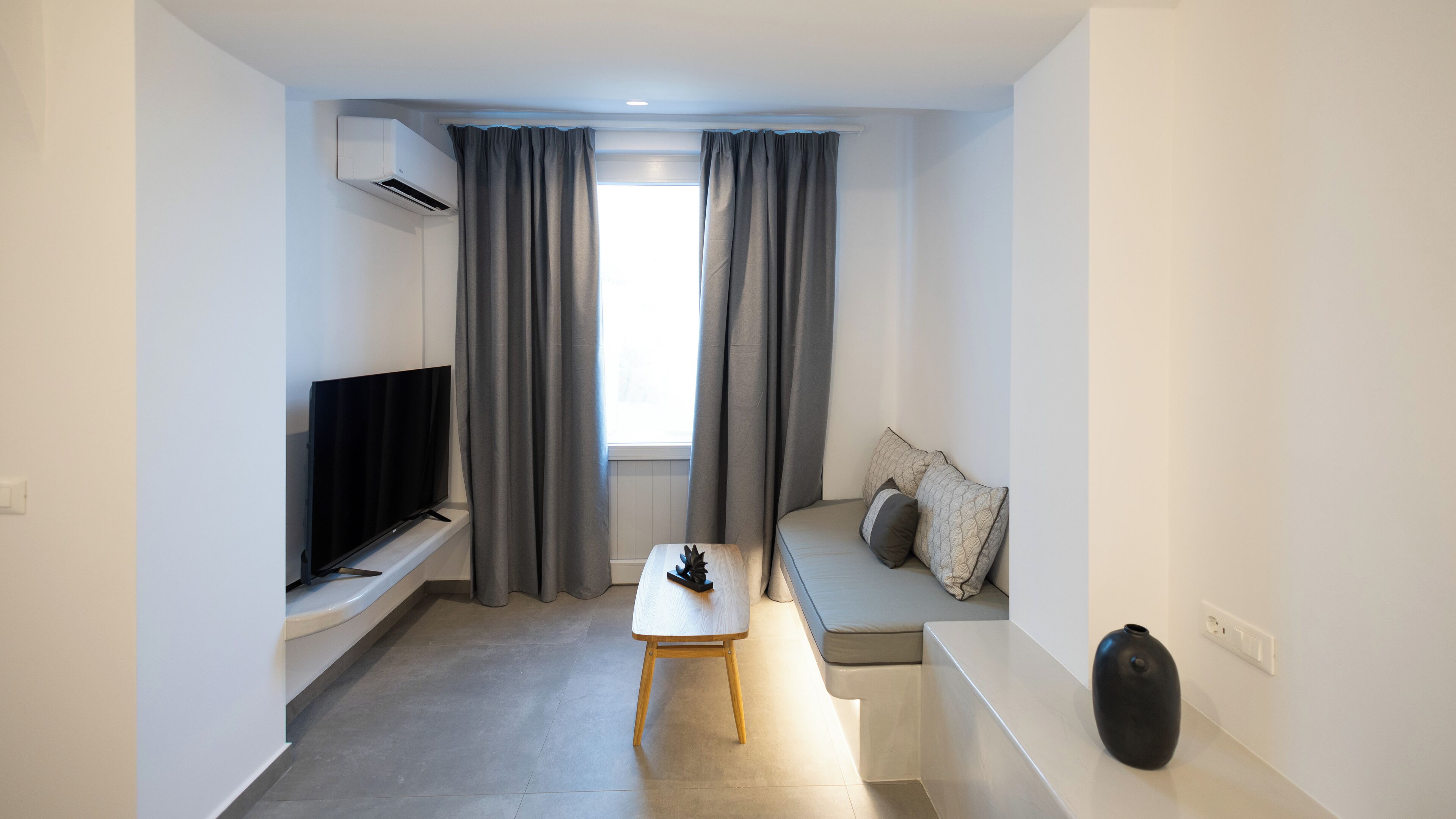 Ein modernes Wohnzimmer mit Flachbildfernseher, einem kleinen Tisch, einer Couch mit Kissen und einem großen Fenster mit Vorhängen.