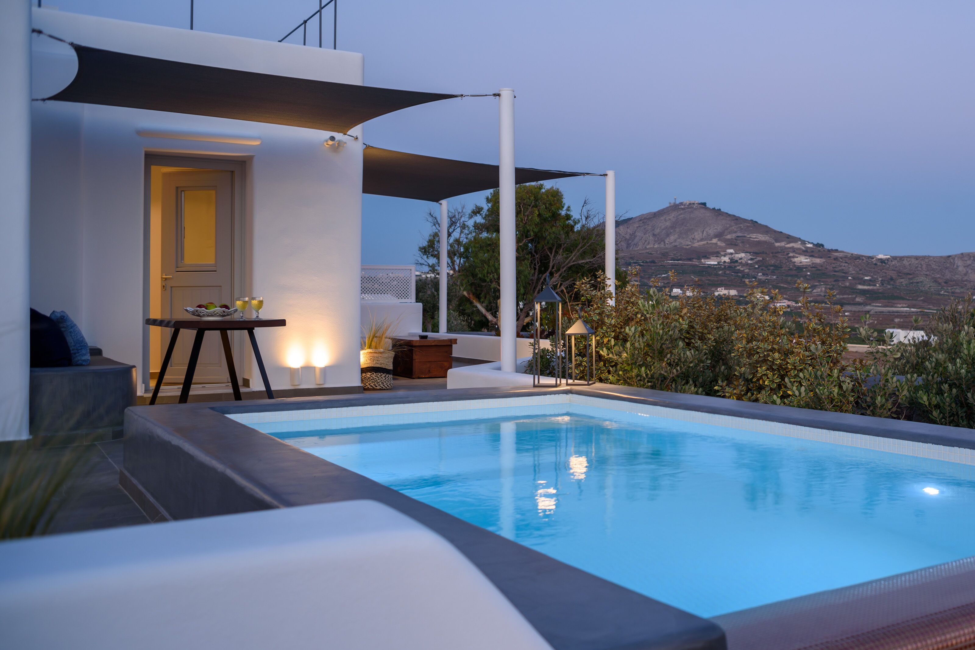 A modern outdoor pool area with a view of a mountain, a door, and a table with a bowl of fruit and glasses.