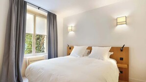 A neatly made bed with white linens, a window with curtains, and two wall-mounted light fixtures.