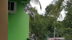 Uma varanda com telhado verde, chão de azulejos, uma cadeira de plástico azul e guarda-corpo de metal.