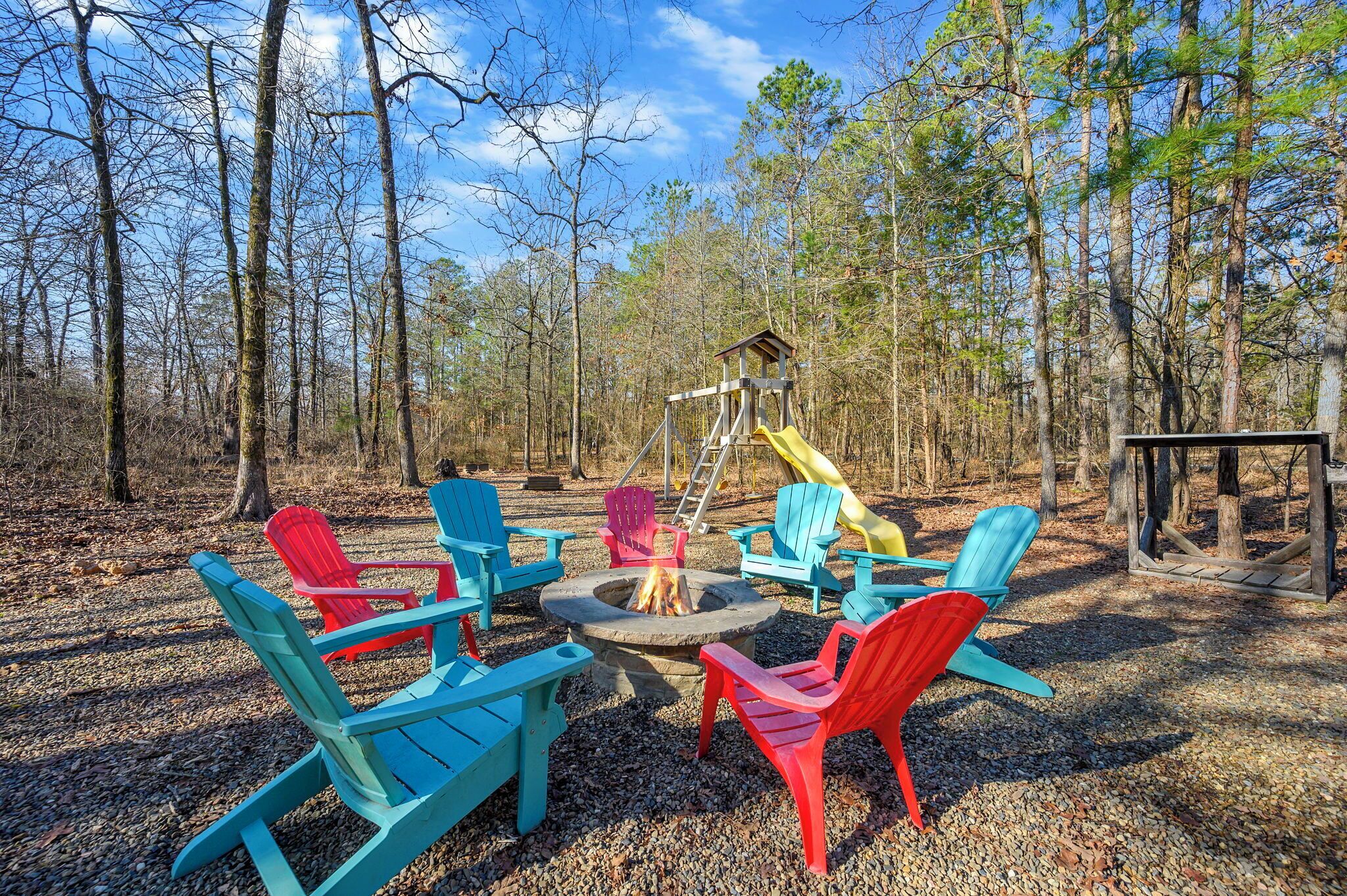 A forested area with a fire pit, colorful Adirondack chairs, and a playground structure.