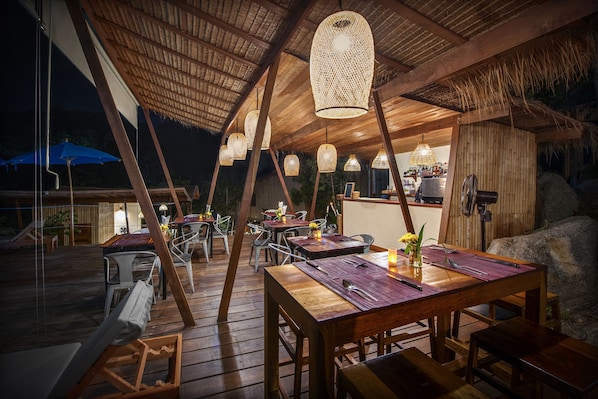 An outdoor dining area with wooden tables and chairs, hanging lanterns, and a thatched roof structure.