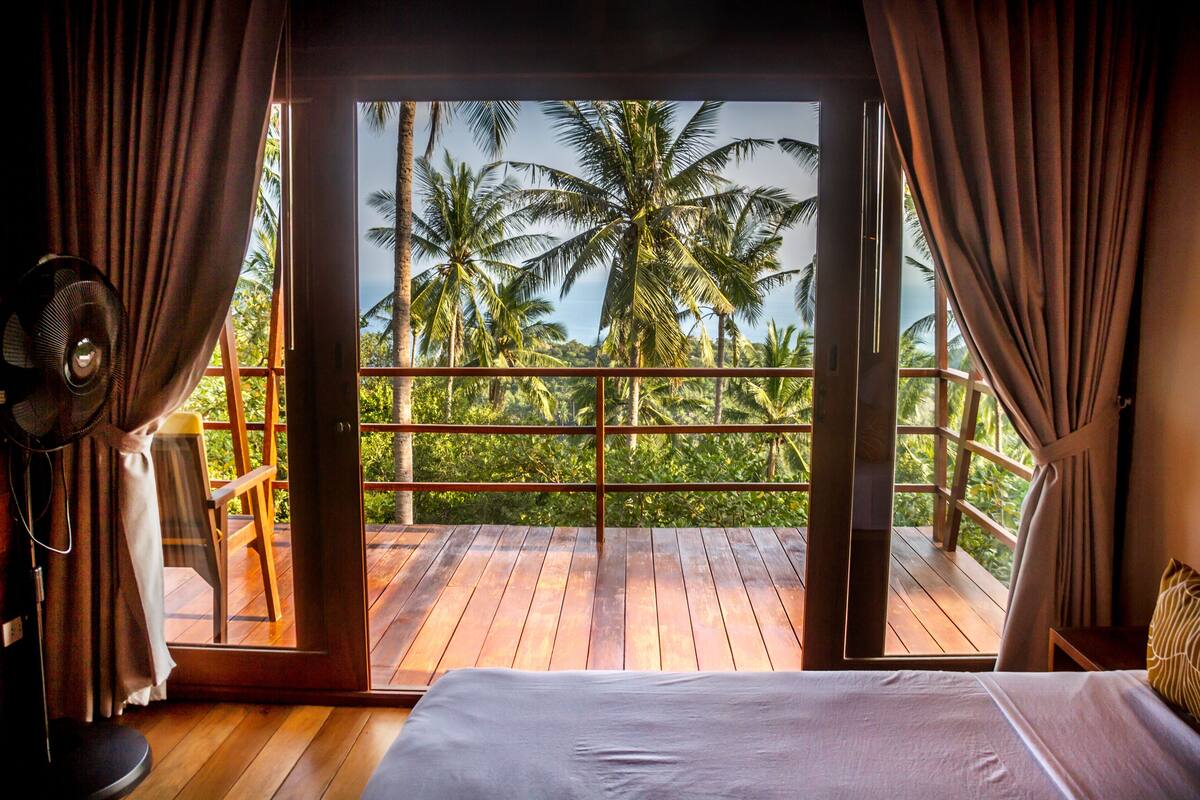 A bedroom with a wooden floor, a bed, a fan, and a view of palm trees through a balcony.