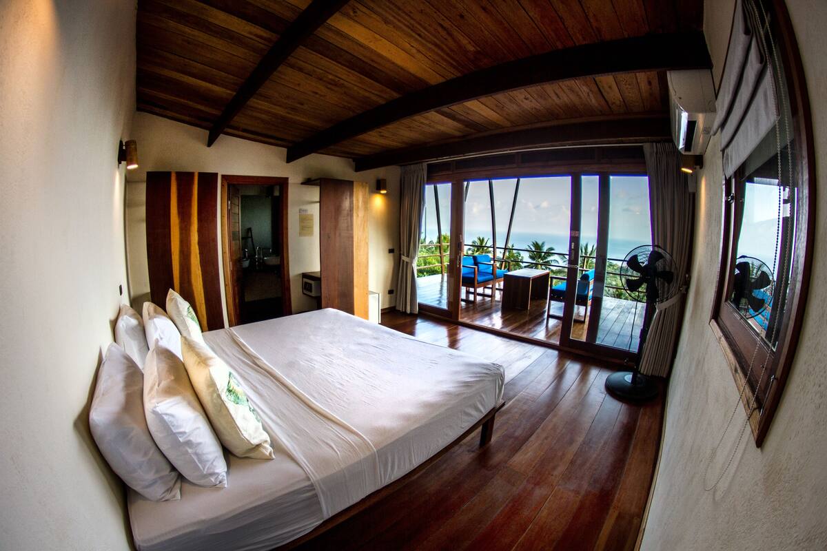 A bedroom with a large bed, wooden flooring, a view of the ocean, and a balcony with a table and chairs.