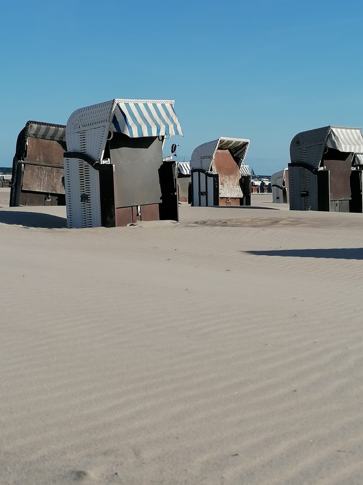 A row of simple, utilitarian beach huts with corrugated metal roofs and walls, situated on a sandy beach under a clear sky.