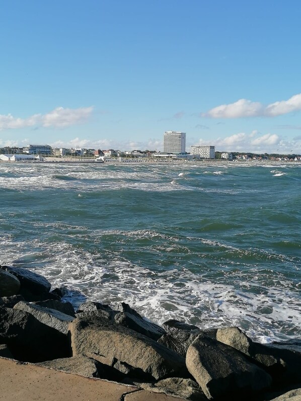 Vista costera con olas rompiendo contra rocas, un muelle y edificios al fondo.