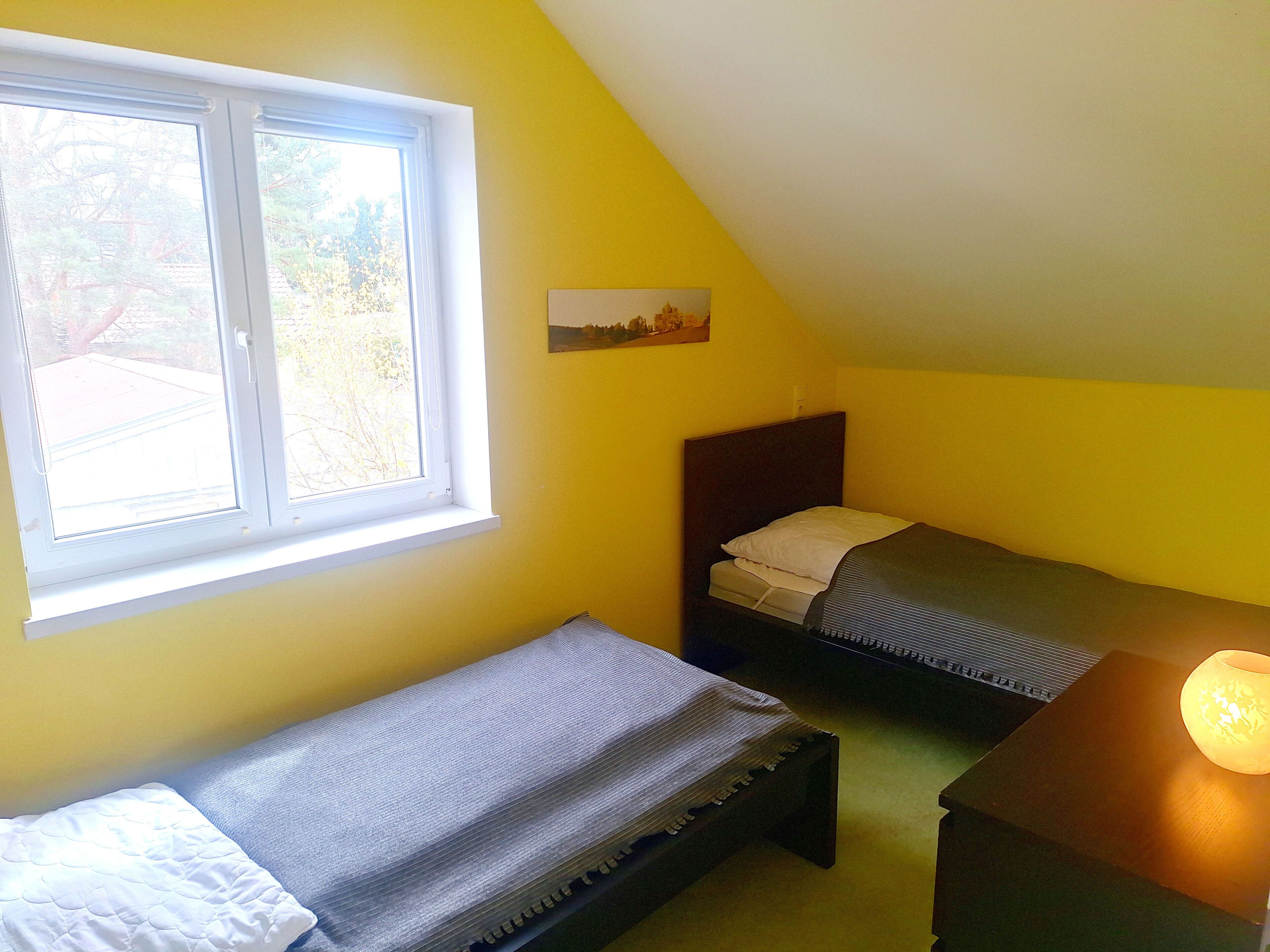 Eine kleine, einfache Unterkunft mit einem Fenster, einem Bett, einem Nachttisch und einer Lampe.