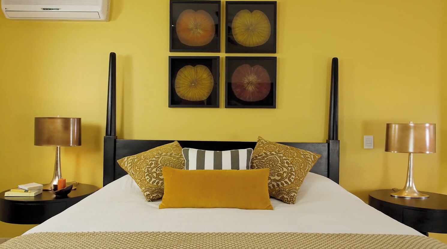 Habitación de hotel con cama, dos mesitas de noche, aire acondicionado de pared y cuatro cuadros enmarcados sobre la cama.