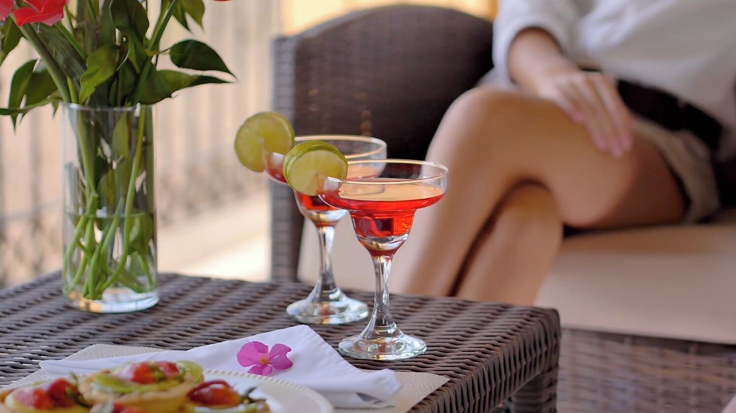 Et flettet bord med to martini-glas fyldt med røde cocktails, en vase med grønne bønner og en tallerken med mad.