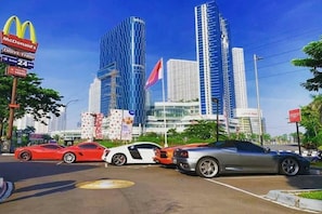 Een parkeerplaats met luxe sportauto's, een McDonald's bord en moderne hoge gebouwen op de achtergrond.