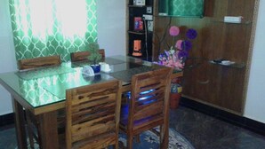 A dining area with a glass-top table, wooden chairs, a small potted plant, and a decorative vase with flowers. - Maria's House  (Shiroda)
