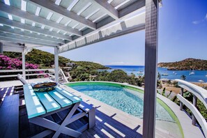 Una piscina con vista al mar y a embarcaciones, una terraza de madera con mesa y sillas, y una pérgola.
