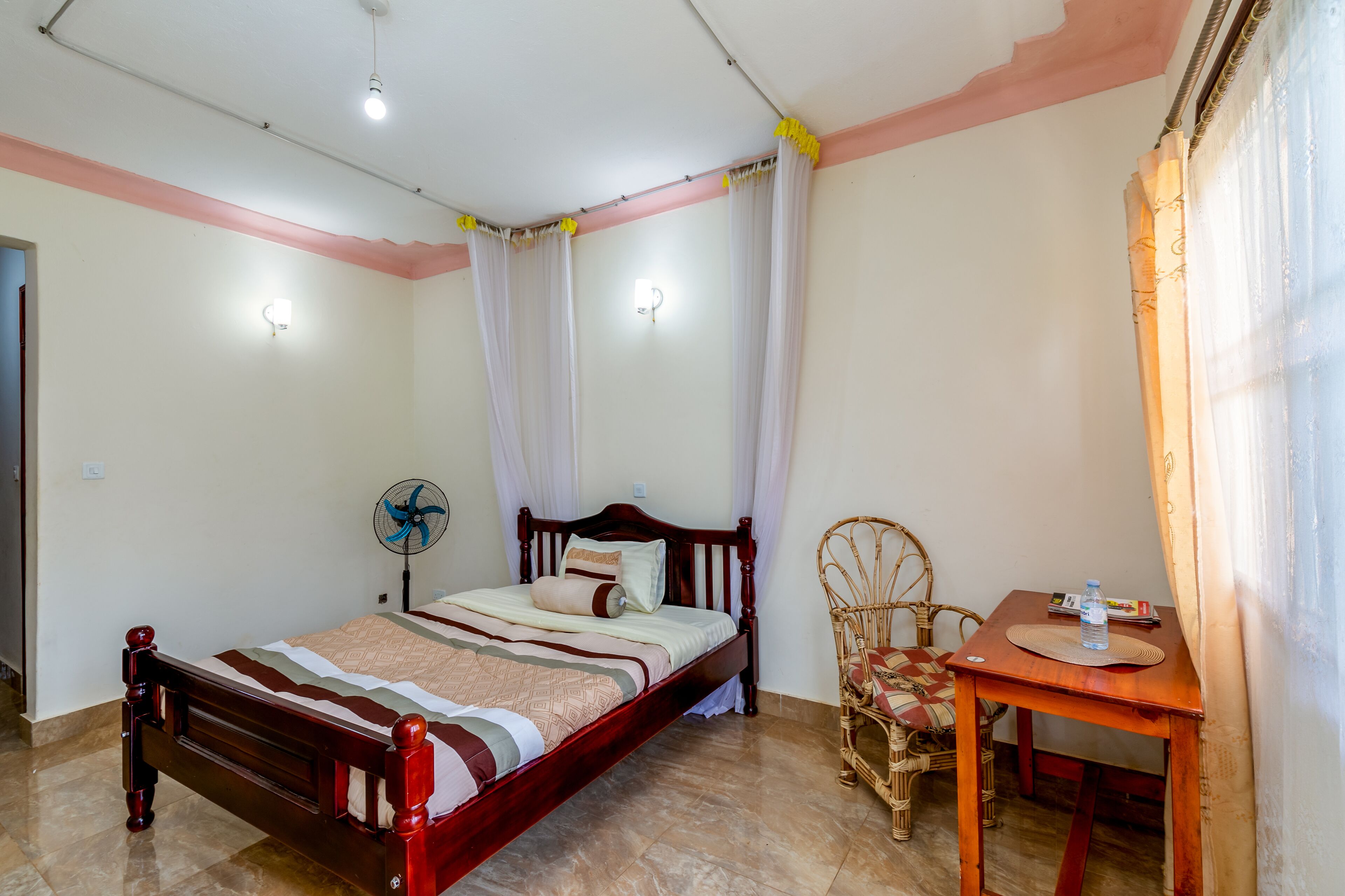 Quarto bem arrumado com cama de madeira, uma mesinha com uma garrafa de água, um ventilador e uma cadeira de vime.