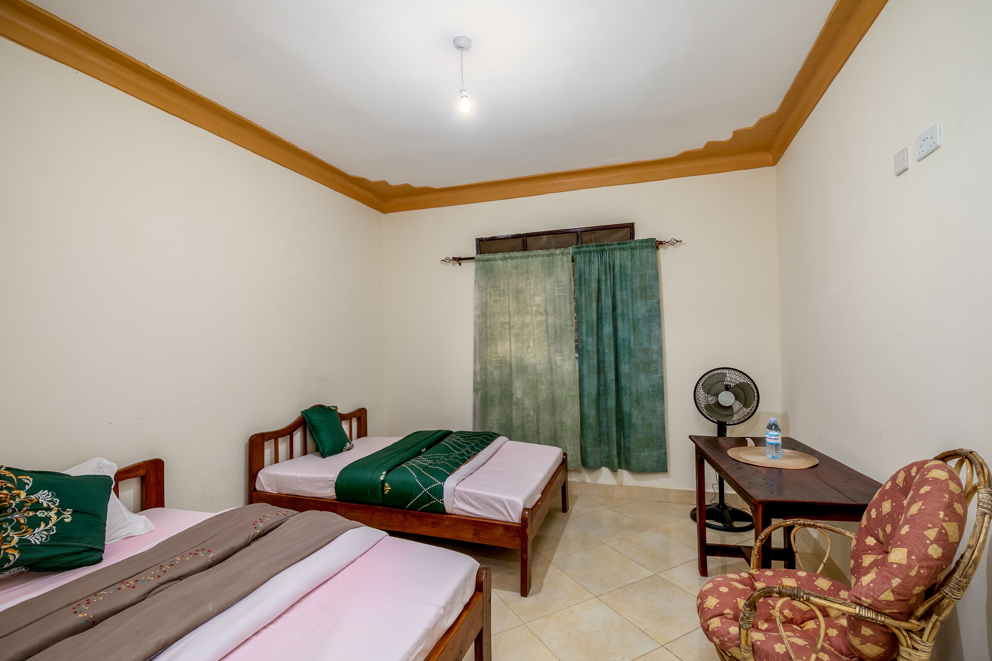 Un dormitorio con dos camas de madera, una silla, una mesa, un ventilador y una ventana con cortinas.
