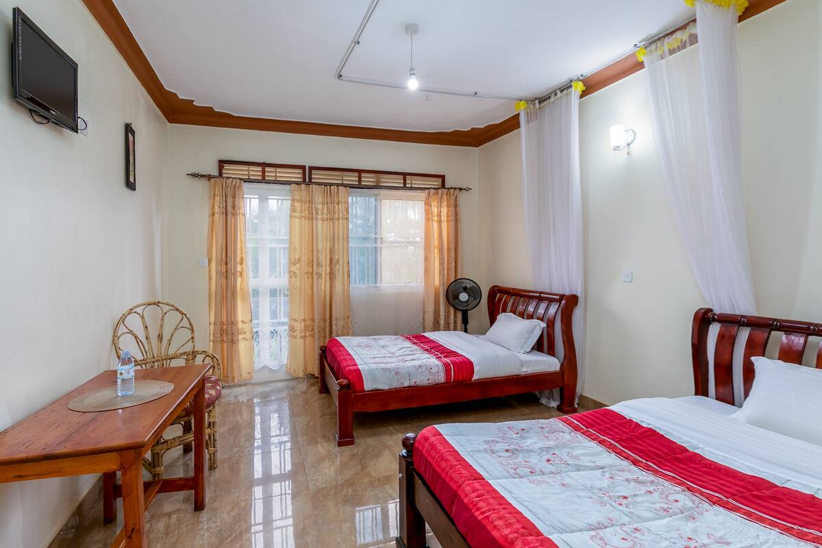 A room with two wooden beds, a wooden table with a chair, a standing fan, and a TV on the wall.