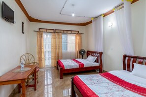Une pièce avec deux lits en bois, une table en bois avec une chaise, un ventilateur sur pied et un téléviseur fixé au mur.