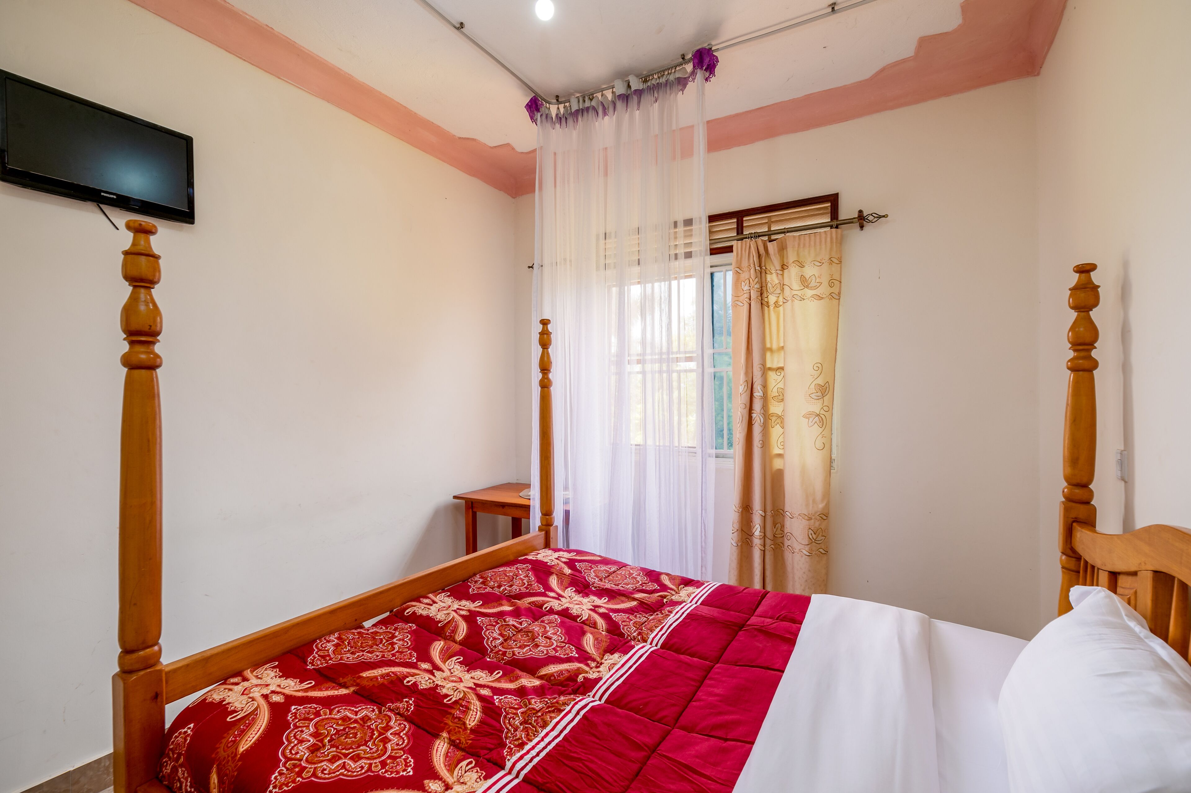 A bedroom with a wooden bed, a TV mounted on the wall, and a window with sheer curtains.