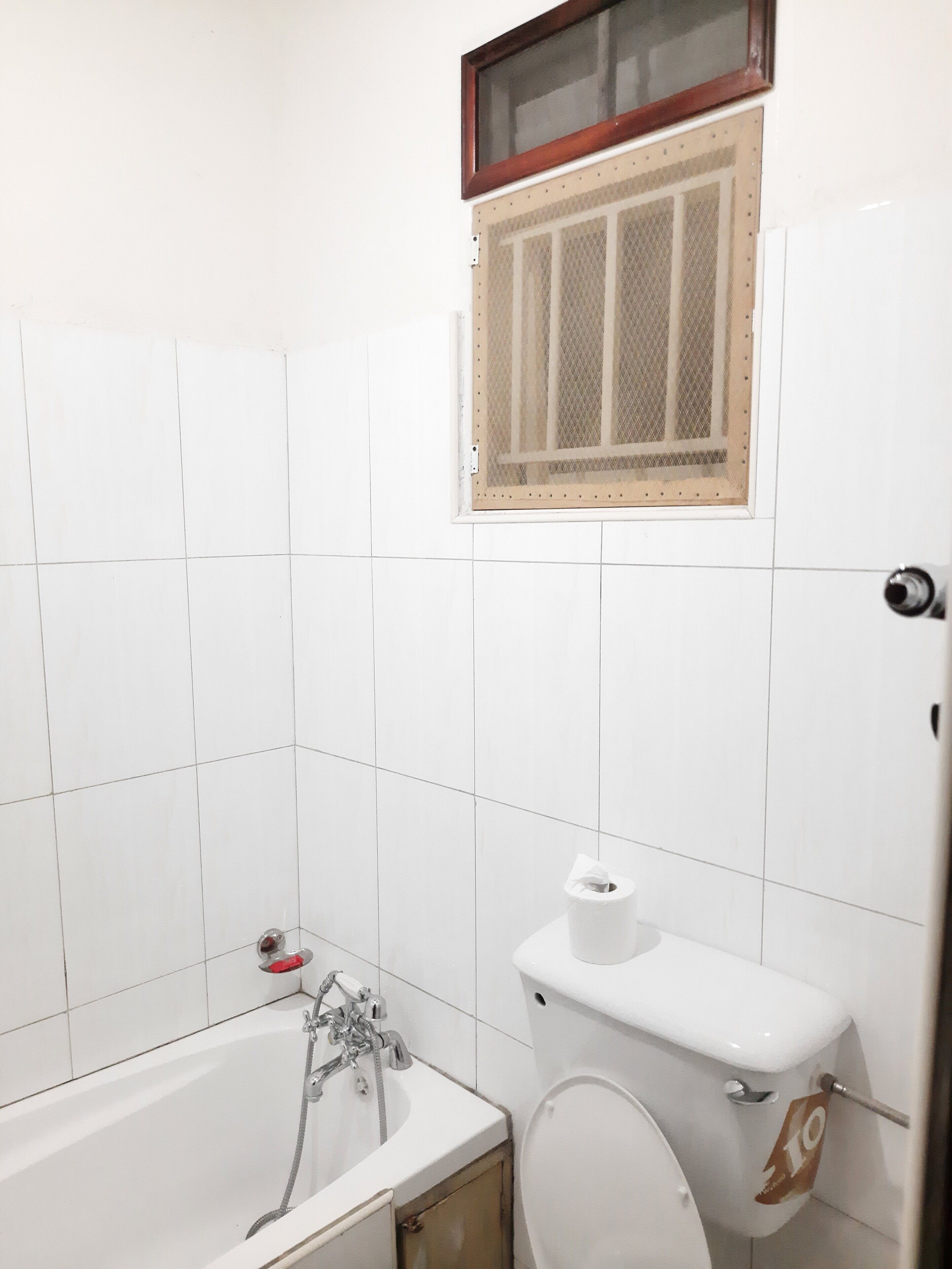 Ein Badezimmer mit weißen Fliesen, einer Badewanne, einer Toilette und einem Fenster mit Insektengitter.
