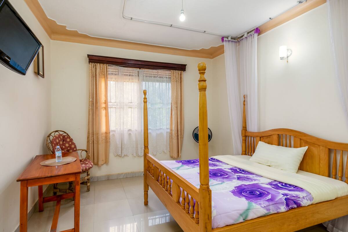 A bedroom with a wooden four-poster bed, a bedside table with a bottle, a chair, and a TV on the wall.