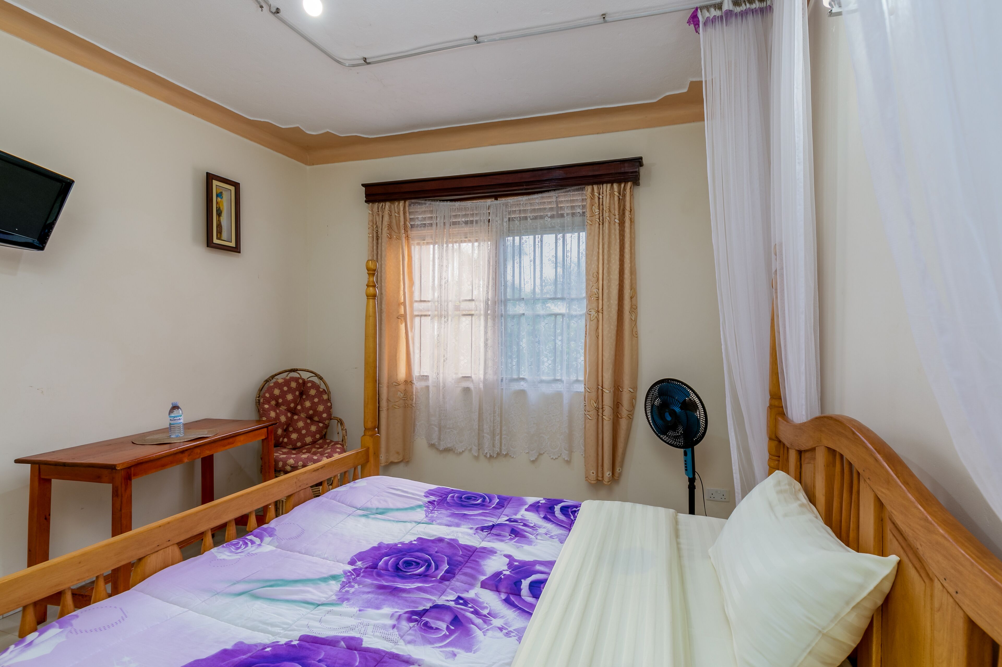 A bedroom with a bed, a wooden headboard, a bedside table with a water bottle, a chair, a television on the wall, and a window with curtains.
