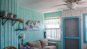 A room with teal walls, a ceiling fan, a beige sofa, a wooden chair, a small table with a lamp, and a window with curtains.