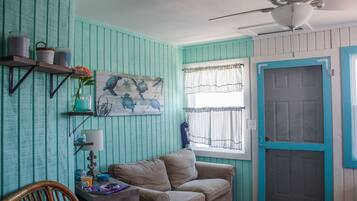 A room with teal walls, a ceiling fan, a beige sofa, a wooden chair, a small table with a lamp, and a window with curtains.