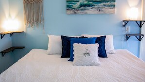 A bed with white and blue bedding, a wall-mounted macrame piece, and a painting of waves.