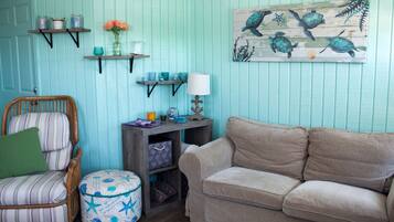 A room with teal walls, a striped chair, a beige sofa, and a wooden side table with a lamp. Shelves with decorative items and a painting of sea turtles are also visible.