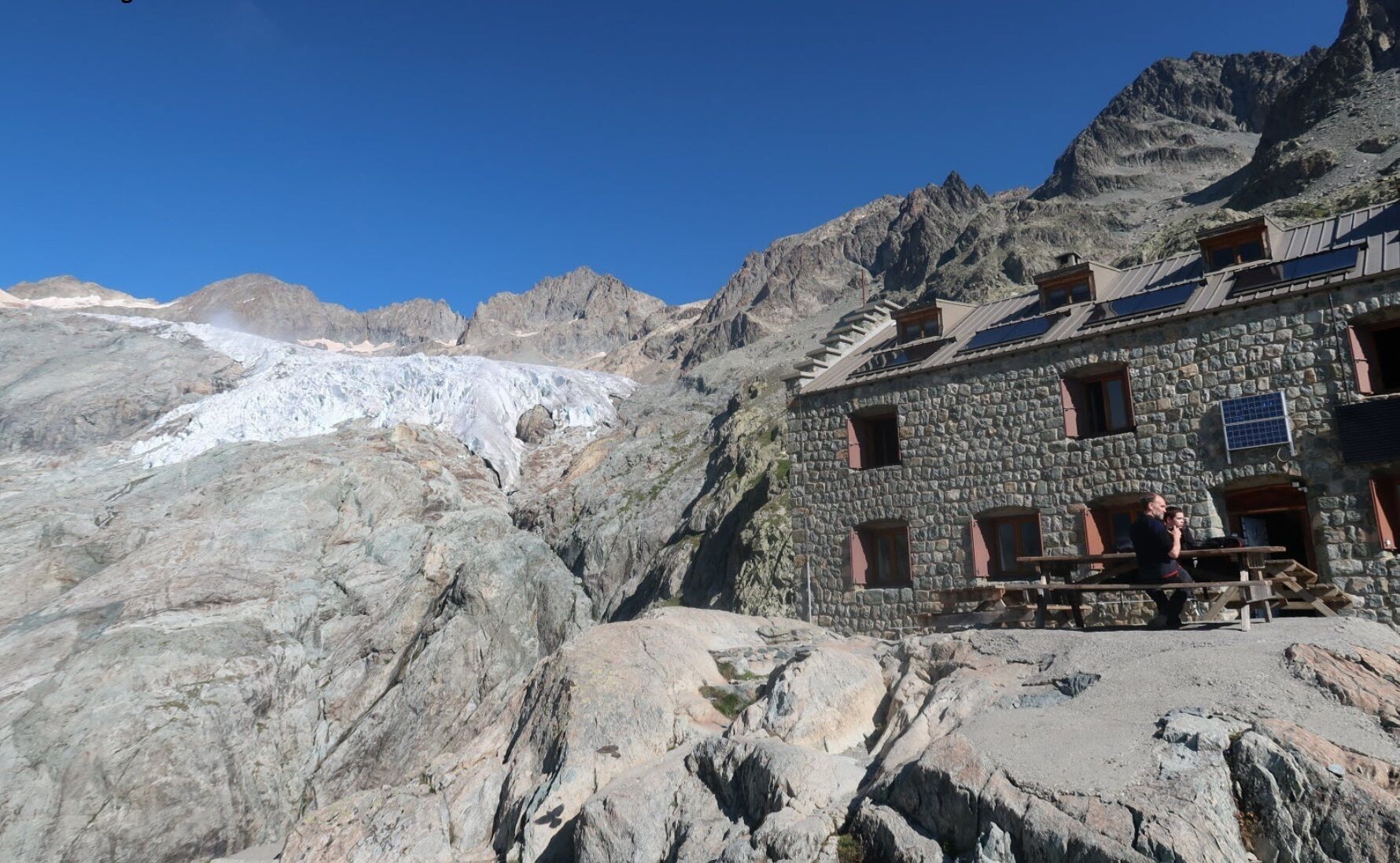 Un edifici de pedra amb teulada inclinada, una banca de fusta a l'exterior i un glaçer al fons.