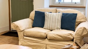 A beige sofa with blue and striped pillows, a wooden table, a woven rug, and a window with a view of buildings. - Cotswold Cottage, with wood burning stove and private courtyard garden (Northleach)