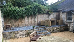 A patio area with stone flooring, a wooden bench, wicker furniture, and a stone wall with climbing plants. - Cotswold Cottage, with wood burning stove and private courtyard garden (Northleach)