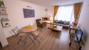 Sala de estar com sofá, mesa de centro e lareira. Há uma mesa de jantar com cadeiras e uma televisão sobre um móvel.