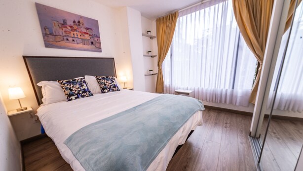 Quarto de hotel com uma cama grande, cabeceira, um quadro na parede, janela com cortinas e mesa de cabeceira com abajur.