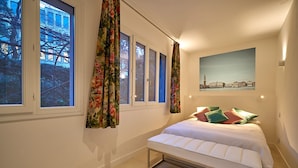 Une chambre à coucher avec un lit, une fenêtre avec des rideaux, un tableau au mur et une vue sur les bâtiments à l’extérieur.