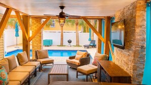 A covered outdoor seating area with a pool, wooden furniture, and a stone wall. - High Tide 5 Bedroom Ocean View House, Heated Pool, Hot Tub, Darts, King beds (Surfside Beach)