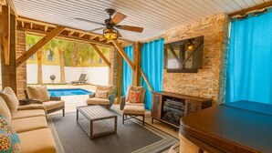A covered outdoor living area with a pool, seating, and a fireplace. - High Tide 5 Bedroom Ocean View House, Heated Pool, Hot Tub, Darts, King beds (Surfside Beach)